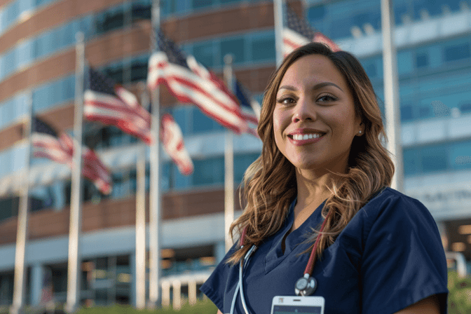 Eine lächelnde Travel Nurse in Uniform steht vor einem amerikanischen Krankenhaus mit mehreren wehenden US-Flaggen im Hintergrund.