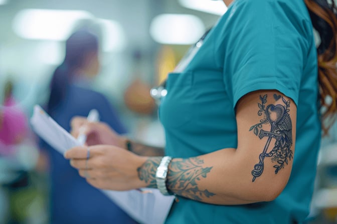 Krankenschwester mit einem detaillierten Tattoo am Arm, trägt türkisfarbene Kasack und schreibt in einer belebten medizinischen Umgebung auf ein Klemmbrett.